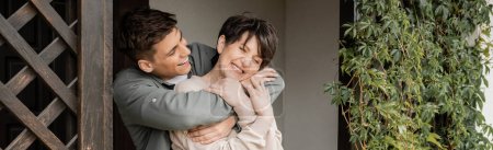 Smiling young man hugging happy middle aged mom while standing near house on porch and celebrating parents day, family traditions and celebrations concept, banner 