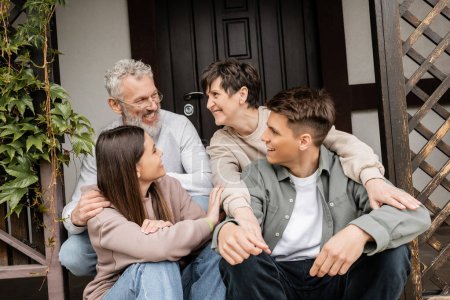 parents d'âge moyen positifs parler tout en embrassant les enfants et assis sur le porche de la maison lors de la célébration de la journée des parents en juin, traditions familiales et concept de célébrations