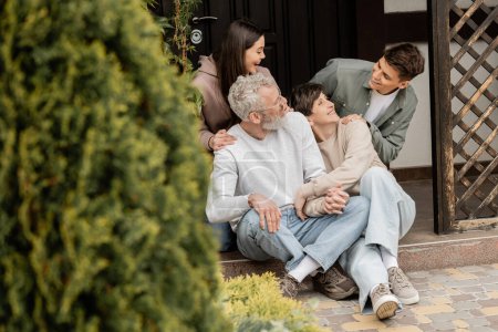 Cheerful kids hugging and talking to middle aged parents while sitting together on porch of house and celebrating parents day in june, family traditions and celebrations concept