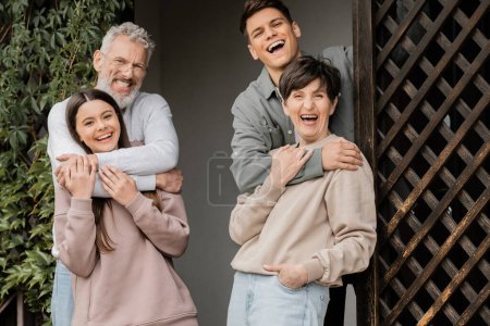 Positive family with kids hugging and looking at camera together while celebrating parents day on porch of house at backyard in june, parent-child relationship concept, special occasion
