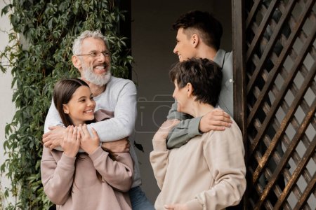 Positiver junger Mann umarmt Mutter mittleren Alters und spricht mit Vater und Schwester im Teenageralter bei Elterntagsfeier, während er auf der Veranda des Hauses steht, Eltern-Kind-Beziehungskonzept