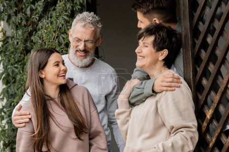 Smiling teenage girl talking to family hugging while celebrating parents day and standing together on porch of house in june, parent-child relationship concept, tradition and celebration