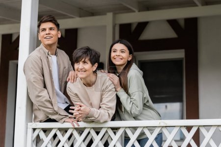 Positive siblings looking away while hugging middle aged mother and celebrating parents day, standing on porch of house in june, parent-child relationship concept, special occasion