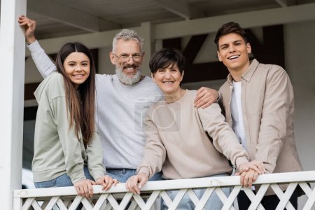 Portrait of positive middle aged parents looking at camera while standing near kids and celebrating parents day near house on porch, parent-child relationship concept, special occasion