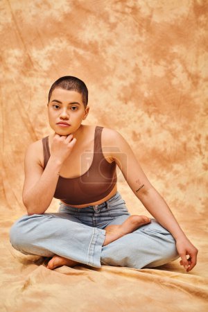 body confidence, acceptance, curvy young and tattooed woman in jeans and crop top sitting with crossed legs on mottled beige background, personal style, self-acceptance, generation z, denim fashion