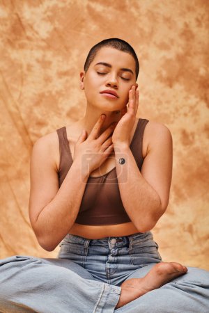 body image, relaxation, curvy young and tattooed woman in jeans and crop top sitting with crossed legs on mottled beige background, closed eyes, personal style, self-acceptance, generation z 