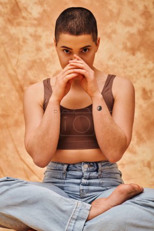 body positivity, curvy and tattooed woman in jeans and crop top sitting with crossed legs on mottled beige background, looking at camera, personal style, self-acceptance, generation z 