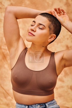 Körperpositivität, kurzhaarige, kurvige und tätowierte Frau im bauchfreien Top posiert auf fleckigem beigem Hintergrund, geschlossene Augen, Körperliebe, Selbstakzeptanz, Generation z, Hände in Nackennähe, kurzhaarig 