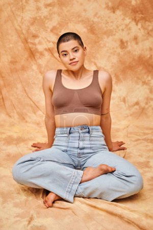 body positivity, casual attire, curvy young and tattooed woman in jeans and crop top sitting with crossed legs on mottled beige background, personal style, self-acceptance, generation z 