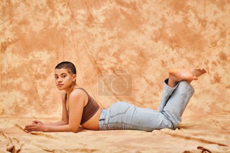 Photo for Body positivity, casual attire, curvy young and tattooed woman in jeans and crop top lying on mottled beige background, looing at camera, denim fashion, personal style, self-acceptance, generation z - Royalty Free Image