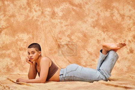 Körperpositivität, Darstellung des Körpers, kurvige junge tätowierte Frau in Jeans und bauchfreiem Oberteil auf fleckigem beigem Hintergrund liegend, Blicke vor die Kamera, Jeansmode, persönlicher Stil, Generation z 