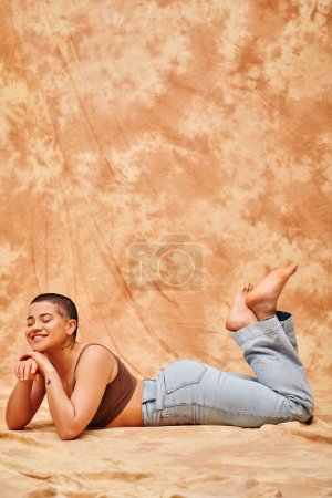 body positivity, representation of body, curvy and tattooed woman in jeans and crop top lying on mottled beige background, smiling with closed eyes, denim fashion, personal style, generation z 