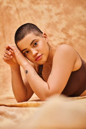 body positivity, curvy and tattooed woman in crop top lying on mottled beige background, looking at camera, representation of body, different body shapes, generation z, youth, blurred foreground 