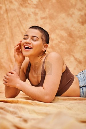 body positivity, curvy and excited woman in crop top posing with hand near face on mottled beige background, tattooed, representation of body, different body shapes, generation z, youth 