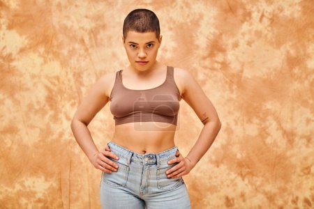 body positivity movement, curvy young woman with tattoos posing in jeans and crop top on mottled beige background, hands on hips, representation of body, confidence, casual attire, generation z 