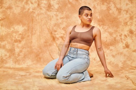 Photo for Body positivity, denim fashion, curvy and tattooed woman in jeans and crop top sitting on mottled beige background, casual attire, looking away, self-acceptance, generation z, body diversity - Royalty Free Image