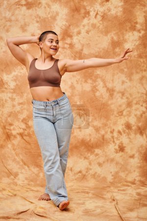 Photo for Body positivity, jeans look, curvy and happy woman in crop top posing with outstretched hand on mottled beige background, casual attire, looking away, self-acceptance, generation z, tattooed - Royalty Free Image