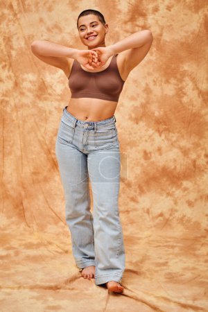 body positivity movement, jeans look, curvy and joyful woman in crop top posing on mottled beige background, casual attire, self-acceptance, generation z, tattooed, smile, full length 
