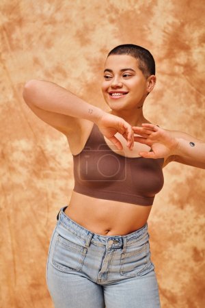 body positivity, joyful and curvy woman in crop top and jeans posing on mottled beige background, yoga pose, self-acceptance, generation z, tattooed, smile, looking away, denim fashion 