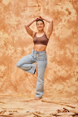 Körperliche Positivität, selbstbewusste, kurvige und lebensfrohe Frau in bauchfreiem Top und Jeans, die auf fleckigem beigem Hintergrund posiert, Yoga-Pose, Selbstakzeptanz, Generation z, tätowiert, Lächeln, volle Länge, Denimmode 