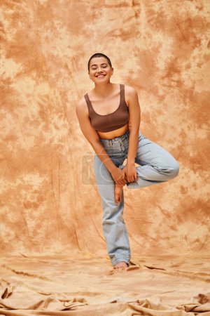 body positivity and confidence, happy young woman in crop top and jeans posing on mottled beige background, casual attire, self-acceptance, generation z, tattooed, smile, full length, denim fashion 