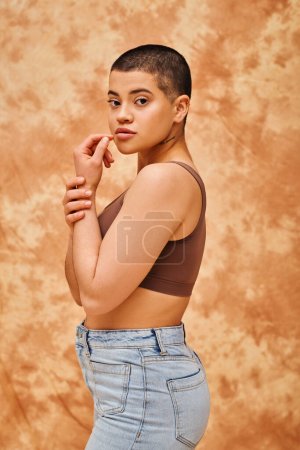 representation of body, curvy and young woman in crop top and jeans posing on mottled beige background, short haired, self-acceptance, generation z, tattooed, different shapes, looking at camera