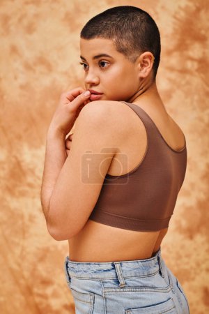 representation of body, curvy and young woman in crop top and jeans posing with hand near lips on mottled beige background, short haired, self-acceptance, generation z, tattooed, different shapes 