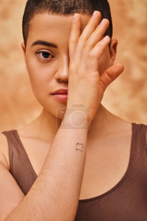 natural look, tattooed young woman with short hair posing on mottled beige background, hand near face, individuality, modern generation z, beauty and confidence, body positivity 