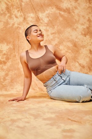 Photo for Body positivity, denim fashion, curvy and pleased woman in jeans and crop top sitting on mottled beige background, tattooed, casual attire, self-acceptance, generation z, body diversity - Royalty Free Image