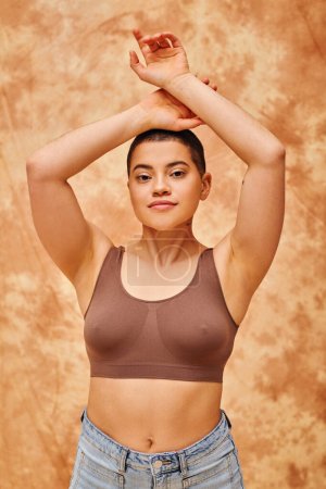 Photo for Body positivity, curvy young woman in crop top posing with raised hands on mottled beige background, looking at camera, representation of body, shapes, generation z, youth, tattooed - Royalty Free Image