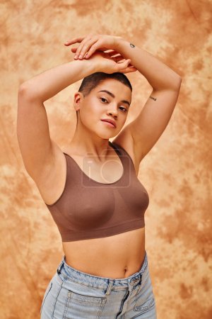 body love, curvy young woman in crop top posing with raised hands on mottled beige background, looking at camera, representation of body, different shapes, generation z, youth, tattooed  