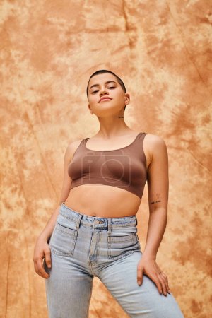 body confidence, jeans look, curvy and tattooed woman in casual attire standing on mottled beige background, looking at camera, self-acceptance, generation z, body diversity, short haired model 