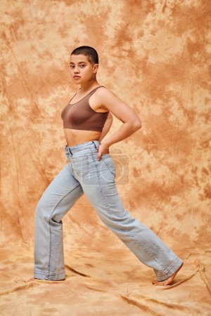 Jeansmode, Generation Z, junge kurvige Frau mit Tätowierungen posiert mit den Händen in den Taschen auf fleckigem beigen Hintergrund, verschiedene Formen, Body Positivity Bewegung, Selbstwertgefühl, Selbstvertrauen