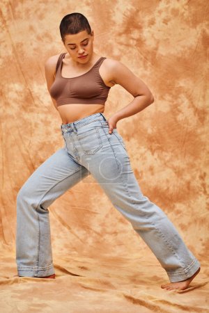 denim fashion, generation z, young curvy woman with tattoos posing with hand in pocket on mottled beige background, different shapes, body positivity movement, self-esteem, confidence, short haired 