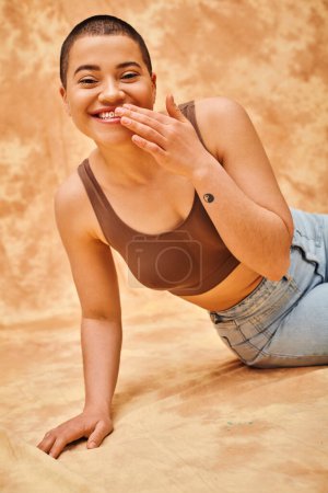 Foto de Moda de mezclilla, gen z, mujer curvilínea alegre con tatuaje sentado sobre fondo beige moteado, movimiento de positividad corporal, autoestima, confianza, modelo de pelo corto, cultura juvenil, felicidad - Imagen libre de derechos