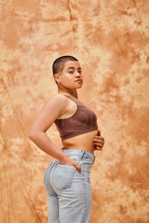 Photo for Denim fashion, gen z, young curvy woman posing on mottled beige background, different shapes, body positivity movement, self-esteem, confidence, short haired model, hand in pocket - Royalty Free Image