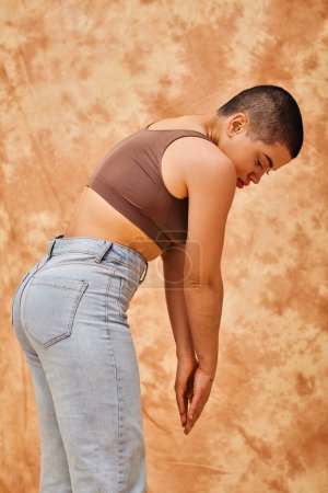 Körperpositivität, Jeansmode, kurvige und tätowierte Frau in Jeans und bauchfreiem Oberteil auf fleckigem beigem Hintergrund, lässige Kleidung Jeansmode, Selbstakzeptanz, Generation z, Körpervielfalt 