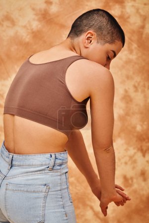 Körpervielfalt, Jeansmode, kurvige und tätowierte Frau in Jeans und bauchfreiem Oberteil auf fleckigem beigem Hintergrund, lässige Bekleidung Jeansmode, Selbstakzeptanz, Generation z, Körperliebe