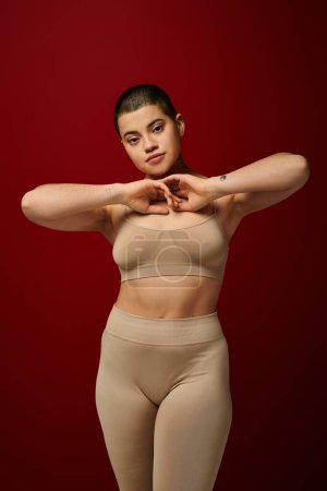 body acceptance, young tattooed woman in beige underwear posing on red background, body positivity, curvy fashion, comfortable in skin, curvy model, generation z, self love, looking at camera 