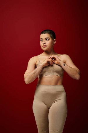 Body Empowerment, junge tätowierte Frau in beiger Unterwäsche posiert auf rotem Hintergrund, Körperpositivität, kurvige Mode, bequem in der Haut, kurviges Modell, Generation z, Selbstliebe, Wegschauen
