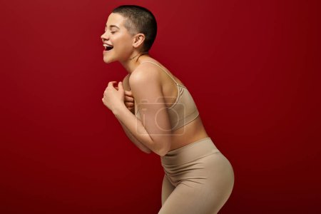 Foto de Autoestima, mujer feliz y tatuada en ropa interior beige posando sobre fondo rojo, moda curvilínea, cómoda en piel, positividad corporal, generación z, diversidad corporal, risa, alegría - Imagen libre de derechos