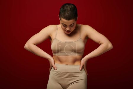 body positive, self-esteem, young woman with short hair and tattoo posing with hands on hips on burgundy background, dark red, curvy fashion, comfortable in skin, female underwear, fashion model 