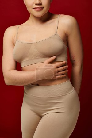 cuerpo positivo, autoestima, vista recortada de mujer joven tatuada posando sobre fondo borgoña, rojo oscuro, moda con curvas, cómodo en la piel, ropa interior femenina, modelo de moda, tipo de cuerpo 