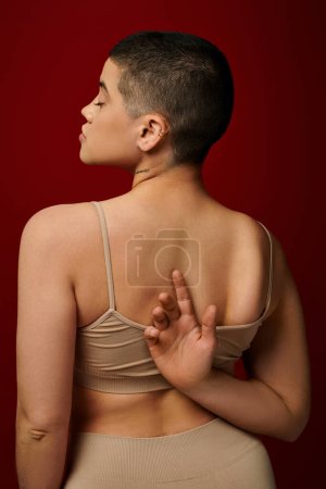 Körper positiv, Selbstwertgefühl, tätowierte junge Frau mit kurzen Haaren und Tätowierung posiert mit der Hand hinter dem Rücken auf weinrotem Hintergrund, dunkelrot, kurvige Mode, angenehm in der Haut, weibliche Unterwäsche 