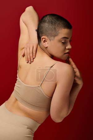 corps positif, confortable dans la peau, jeune femme tatouée avec les cheveux courts et tatouage posant avec la main derrière le dos sur fond bordeaux, rouge foncé, mode courbée, sous-vêtements féminins 