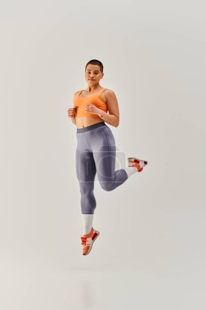 Körperpositivität, junge kurzhaarige Frau springt auf grauem Hintergrund, kurvige Mode, weibliche Fitness, Empowerment, Motivation, Training, Sportbekleidung, Kraft und Gesundheit, Körperbild 