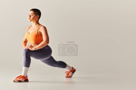 Body Positivity Bewegung, junge kurzhaarige Frau beim Ausfallschritt auf grauem Hintergrund, kurviges Fitnessmodel in Sportbekleidung, Empowerment, Motivation, Training, Kraft und Gesundheit 