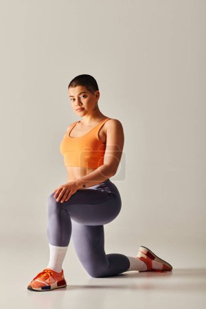 Body Positivity Bewegung, junge kurzhaarige Frau auf Knien vor grauem Hintergrund, kurviges Fitnessmodel in Sportbekleidung, Empowerment, Motivation, Training mit erhobenen Händen, Kraft und Gesundheit 
