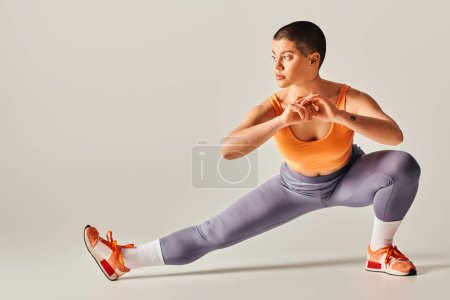 Selbstwertgefühl, Körperpositivität, flexible und kurzhaarige Frau, die Beine vor grauem Hintergrund streckt, kurviges Fitnessmodel in Sportbekleidung, sportlich und selbstbewusst, Empowerment, Motivation 