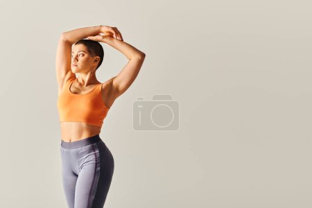 formas del cuerpo, mujer flexible y de pelo corto estiramiento sobre fondo gris, modelo de fitness con curvas en ropa deportiva, atlético y seguro, empoderamiento, motivación, ejercicio con las manos levantadas 
